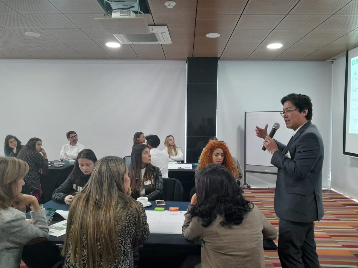 Augusto Ruiz, de pie y con micrófono, expone ante un grupo de asistentes sentados alrededor de mesas de trabajo en una sala empresarial.